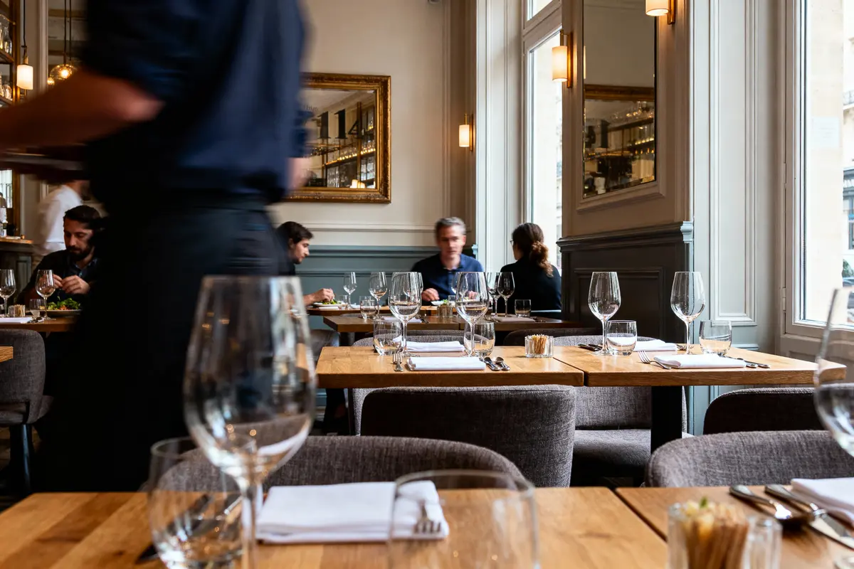 Intérieur de restaurant élégant avec tables en bois, verres à vin, clients et serveurs en mouvement. Ambiance chaleureuse.