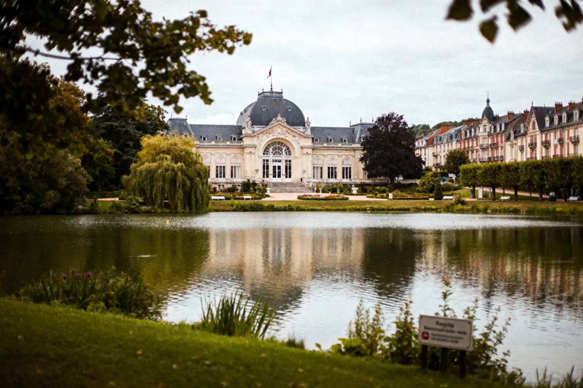Bâtiment historique avec dôme, au bord d'un lac, encadré d'arbres et de pelouses, ciel nuageux.