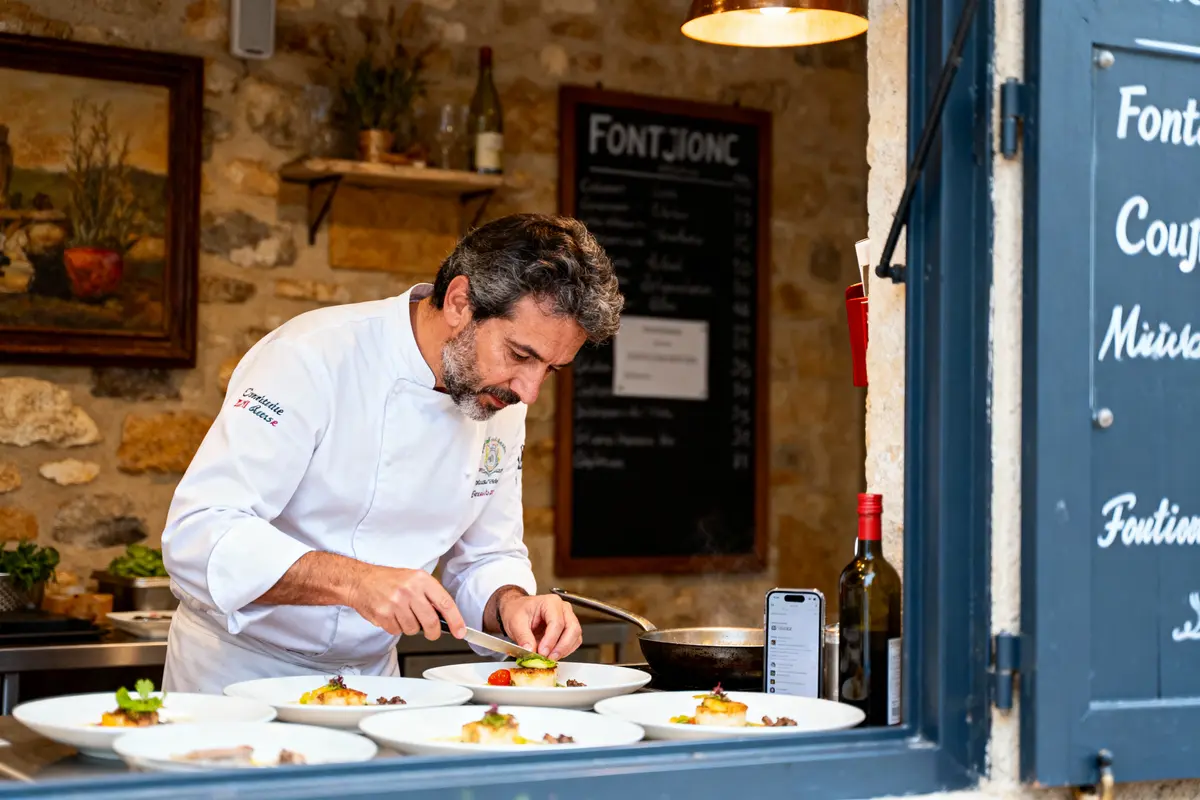 Chef en cuisine décor rustique prépare des plats élégants. Tableaux, étagères, bouteille de vin et smartphone visibles.