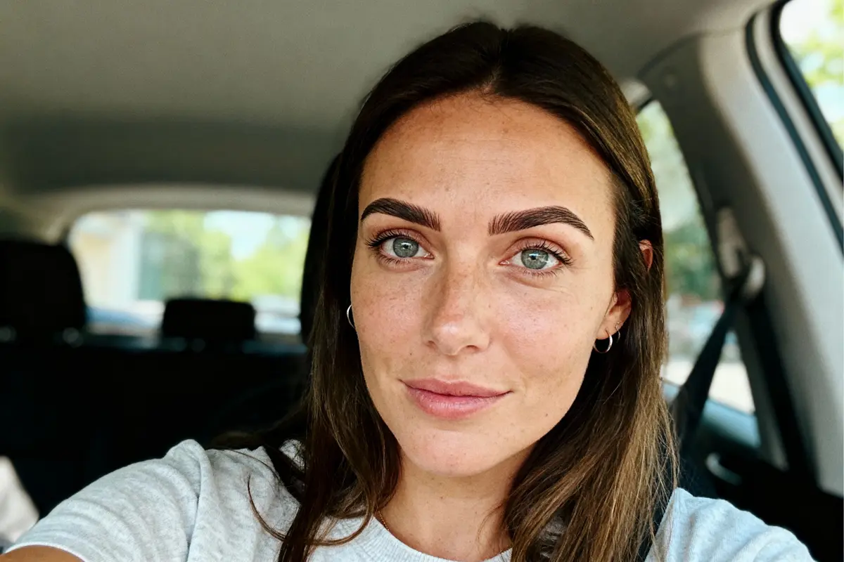Femme aux yeux clairs, cheveux longs, dans une voiture, portant un t-shirt gris, souriant subtilement.