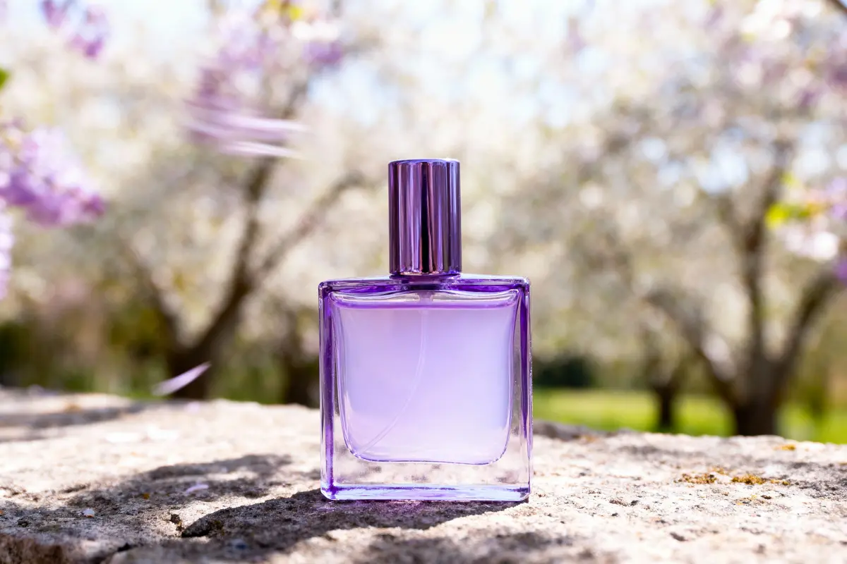 pexels-michal-pech-213601-757318 (1) Bouteille de parfum violette sur pierre, fond flou de nature printanière avec fleurs et arbres.