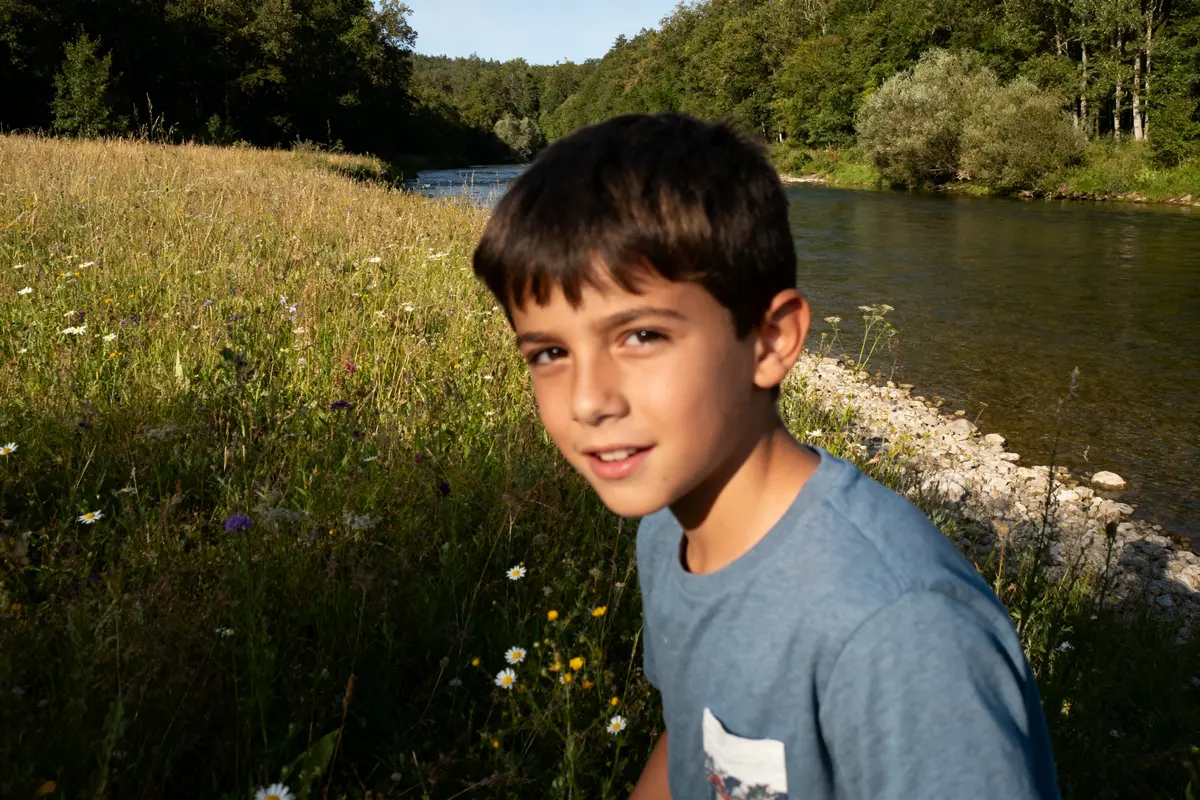 Garçon souriant dans un pré fleuri près d'une rivière; forêt en arrière-plan sous le soleil.