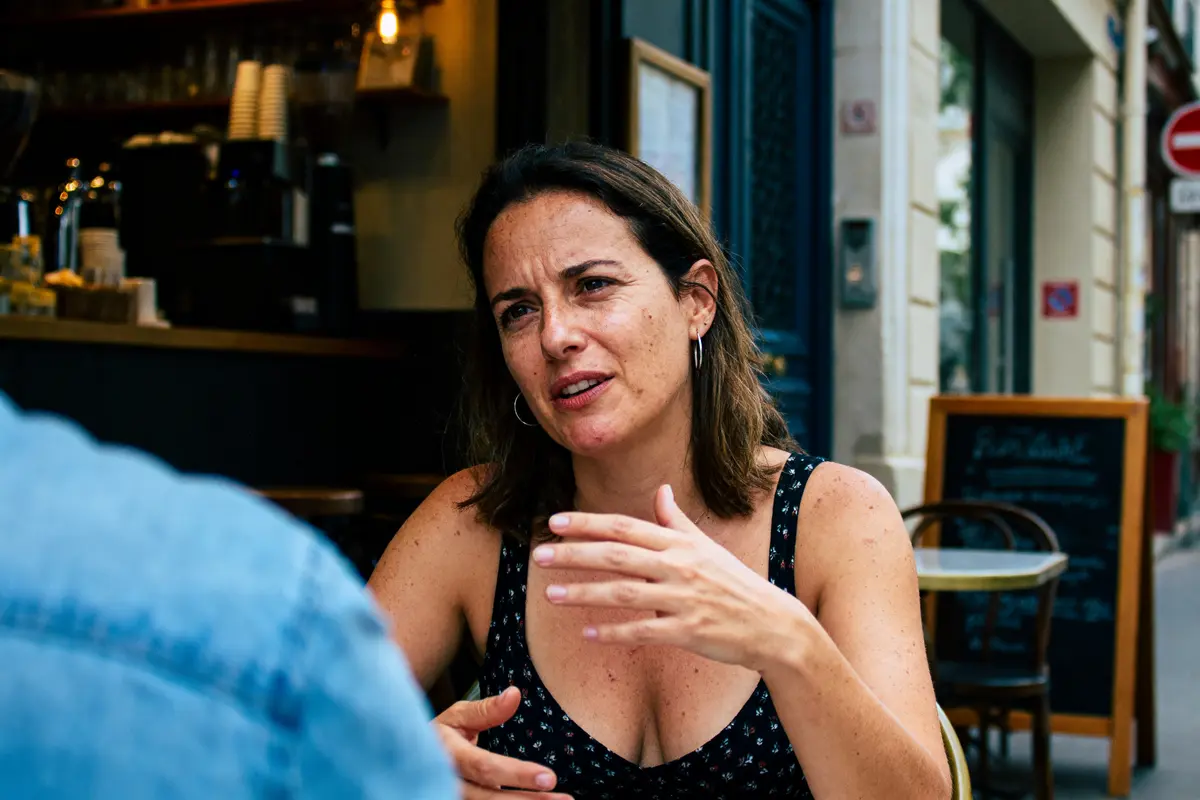 Une femme parlant à l'extérieur d'un café, gesticulant, expression sérieuse, ambiance urbaine.