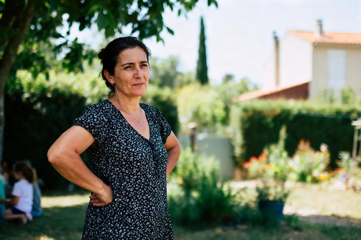 Femme debout dans un jardin ensoleillé, bras croisés, portant une robe à motifs fleuris.