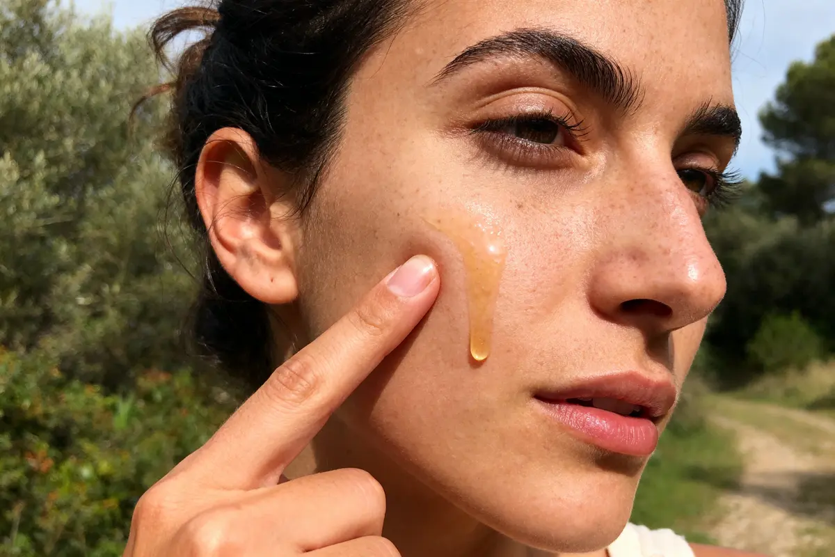 Femme appliquant du gel ou sérum sur sa joue en extérieur, par temps ensoleillé. Peau bronzée.